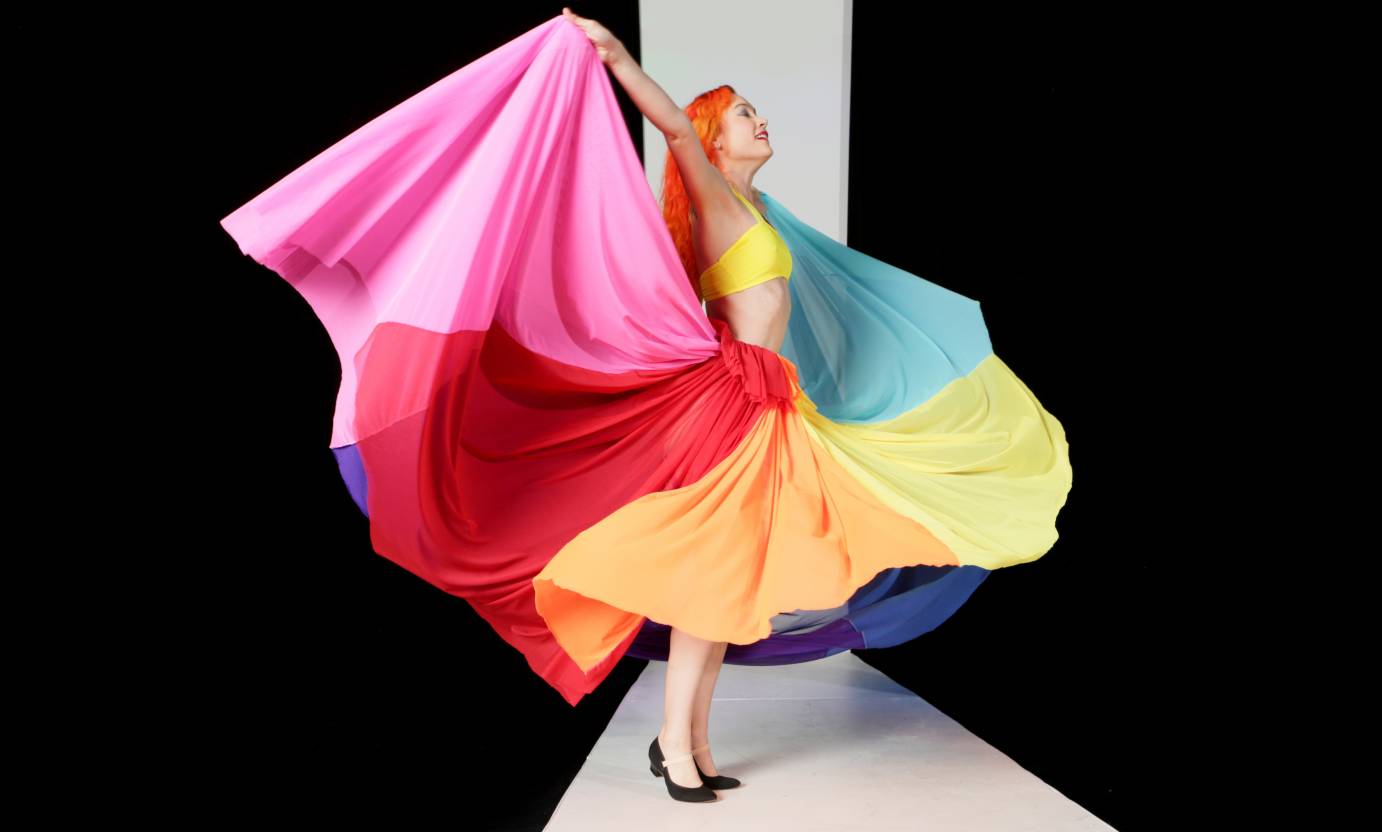A woman with orange hair lifts a rainbow colored skirt off to her sides.